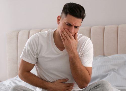 A nauseous man sitting on the bed with his hand over his mouth.